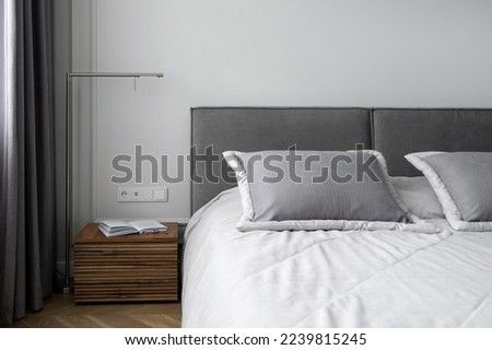 soft pillows and cotton blanket on bed with grey headboard, open book on wooden side table and metal lamp in room with modern interior Royalty-Free Stock Photo #2239815245