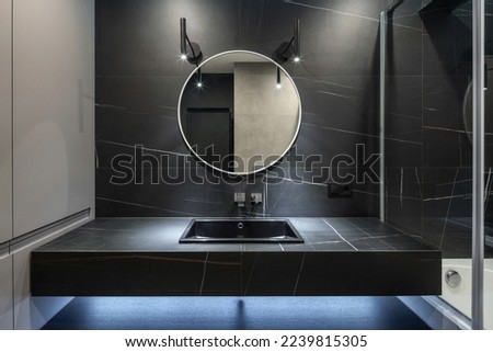 luxury interior in apartment bathroom, stainless steel faucet, black washbowl, metal lamps and round mirror on wall with dark marble tile Royalty-Free Stock Photo #2239815305