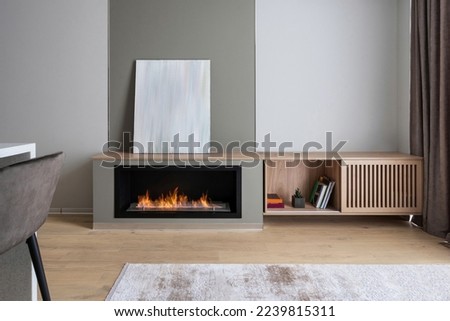 modern interior in living room at stylish apartment, burning flame in electric fireplace, blank art poster, wooden sideboard with books and rug on floor