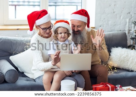 Senior grandparents, attractive grandmother and handsome grandfather doing video call on laptop with cute little granddaughter near decorated Christmas tree. Two generations, family values