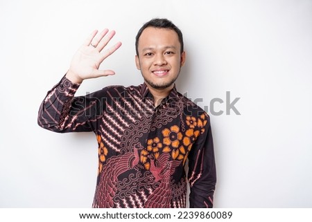 Young cheerful Indonesian man wearing batik shirt with happy positive smile waving hand hello isolated over white color background