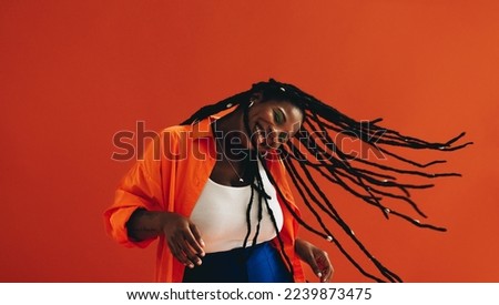 Vibrant young woman flipping her hair playfully while standing in a studio. Excited woman dancing and celebrating against an orange background. Happy young woman having fun with her dreadlocks. Royalty-Free Stock Photo #2239873475
