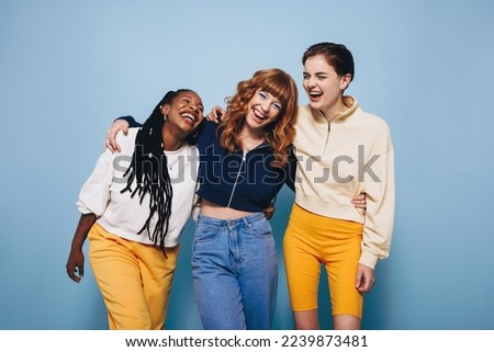 Diverse female friends laughing and having a good time while embracing each other. Group of happy young women enjoying themselves while standing against a blue background. Royalty-Free Stock Photo #2239873481