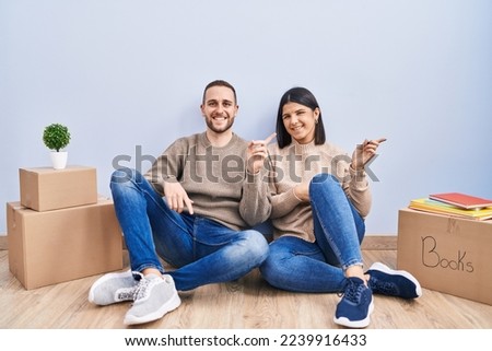 Young couple moving to a new home with a big smile on face, pointing with hand finger to the side looking at the camera. 