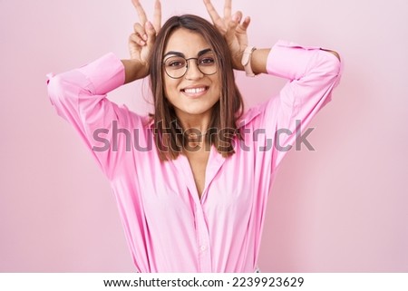 Young hispanic woman wearing glasses standing over pink background posing funny and crazy with fingers on head as bunny ears, smiling cheerful 