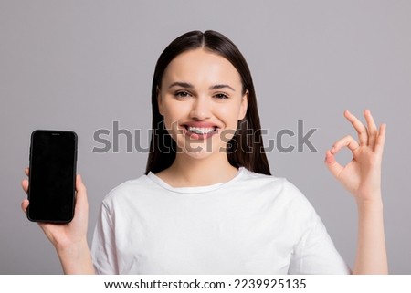 Portrait close up shot on young woman girl in basic white t-shirt standing on grey background in studio isolated holding new last series smartphone selling something on black screen blank shows okay.