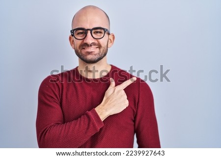 Young bald man with beard standing over white background wearing glasses cheerful with a smile on face pointing with hand and finger up to the side with happy and natural expression 