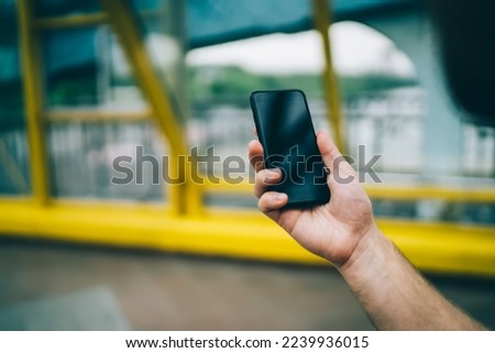 Crop hand of faceless person holding black cellphone with empty screen in glass covered pedestrian bridge on city street  on blurred background