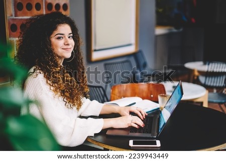 Portrait of female blogger working remotely in cafe interior using laptop computer for share content, skilled student with curly hair looking at camera during freelance time in coffee shop