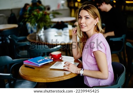 Positive female in casual clothes sitting at wooden table and browsing tablet while resting after workday and looking away in modern cafe Royalty-Free Stock Photo #2239945425