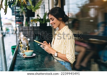 Side view of positive young ethnic female with dark hair in trendy outfit having coffee break with cup of cappuccino and using smartphone on stylish cafe terrace