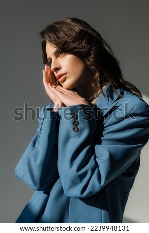 thoughtful brunette woman in blue blazer standing with hands near face and looking away on grey 
