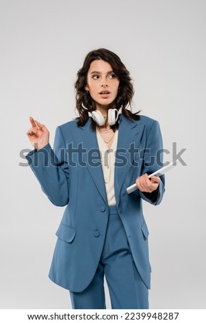 trendy brunette woman with wireless headphones and digital tablet pointing with finger and looking away isolated on grey
