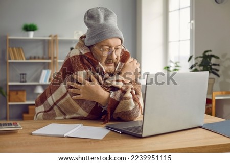 Elderly man in hat sitting at computer wrapped in plaid. Old man freezing at home since broken heating or cold weather winter. Unwell sick pensioneer having fever, shivering listening to online doctor Royalty-Free Stock Photo #2239951115