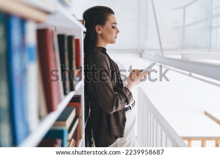 Side view of concentrated young female in casual outfit standing on balcony near bookcase and texting message via cellphone in light modern apartment