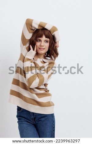 vertical photo of a beautiful, stylish woman in a fashionable striped sweater standing on a light background and relaxed posing with her hands near her face smiling pleasantly
