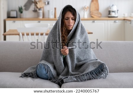 Sick unhappy woman frustrated looking at thermometer having cold during winter walk or work on frost. Diseased sad girl wrapped in blanket has chills and stress sits on sofa in unheated apartment Royalty-Free Stock Photo #2240005767