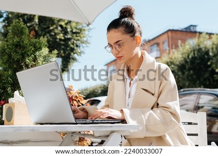 Remote work, freelance. Business stylish woman in glasses working on laptop, concentrated looking at device screen while sitting in street cafe on sunny day. Royalty-Free Stock Photo #2240033007
