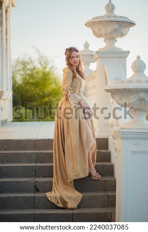 A beautiful young woman in a historic eighteenth century gold dress stands on the stairs of the mansion. Princess in the palace. fabulous image Royalty-Free Stock Photo #2240037085