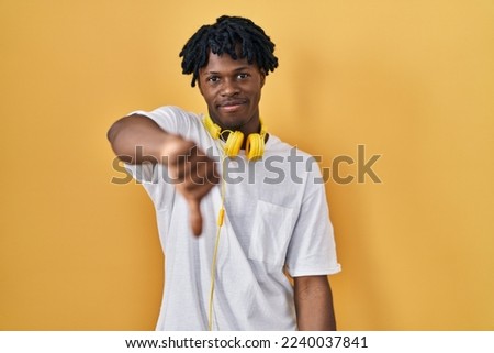 Young african man with dreadlocks standing over yellow background looking unhappy and angry showing rejection and negative with thumbs down gesture. bad expression. 