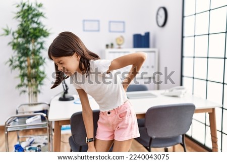 Young hispanic girl standing at pediatrician clinic suffering of backache, touching back with hand, muscular pain  Royalty-Free Stock Photo #2240043075