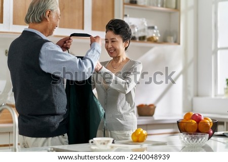 loving senior asian man husband helping wife put on apron in kitchen at home Royalty-Free Stock Photo #2240048939