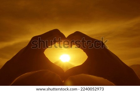 Silhouette of hand making heart sign to the bright sun rising over the Silhouette of mountain peak