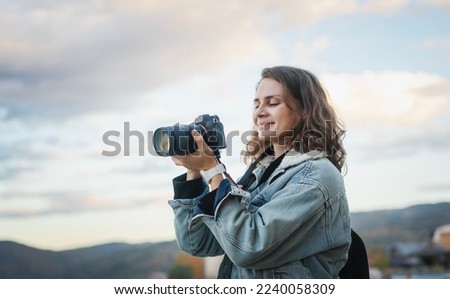 Young cheerful woman traveler in a denim jacket with a backpack holds a camera in her hands while standing on the shore of the mountain  lake Royalty-Free Stock Photo #2240058309