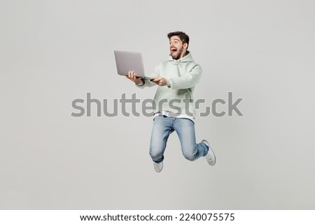 Full body young caucasian happy surprised excited amazed IT man wear mint hoody jump high hold use work on laptop pc computer isolated on plain solid white background studio. People lifestyle concept