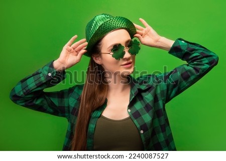 Attractive stylish young woman wearing party green hat, clover shaped glasses, checkered plaid shirt and looking aside isolated on green background.
