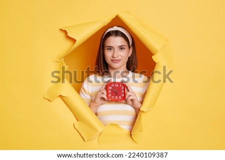 Portrait of pretty young Caucasian woman wearing striped T-shirt and hair band, breaking through paper hole in yellow wall, holding alarm clock, looking at camera with positive expression.