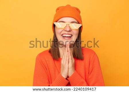 Portrait of pleased hipster woman wearing orange sweater, hat and glasses in minecraft style posing isolated over yellow background, keeps palms in praying gesture, pleading.