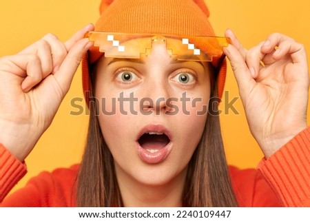 Portrait of shocked hipster woman wearing orange sweater, hat and glasses in minecraft style posing isolated over yellow background, raised eyeglasses and looking with big eyes at camera.