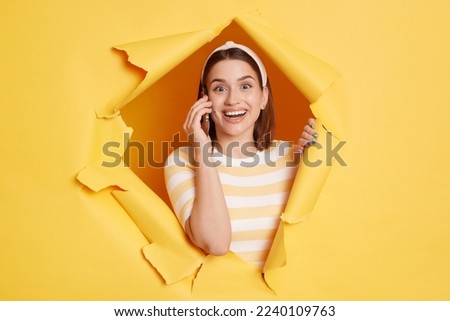 Portrait of smiling amazed excited young woman wearing striped T-shirt and hair band breaking through paper hole in yellow wall, looking at camera, smiling happily.