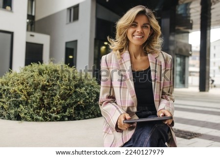 Beautiful young caucasian business woman working on tablet near office. Blonde smiles looking at camera, wears jacket. Concept of use Royalty-Free Stock Photo #2240127979