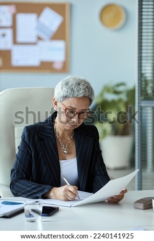 Vertical portrait of senior woman as female boss reading document at workplace in office