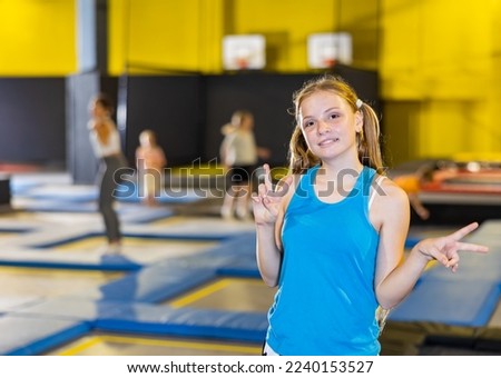 Cheerful confident sporty teen girl enjoying time in indoor trampoline arena, standing with jumping children in background and giving V sign