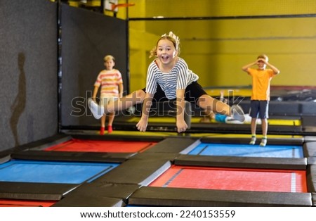 Expressive happy tween girl having fun during free-jumping session in indoor trampoline arena, doing middle split in jump.. Royalty-Free Stock Photo #2240153559