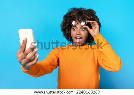 Shocked amazed emotional pretty millennial african american woman with bushy hair looking at modern phone and removing glasses, reading exciting news or ad, blue studio background Royalty-Free Stock Photo #2240163289