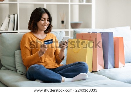 Online Shopping Concept. Happy Arab Female Using Smartphone And Credit Card While Sitting On Couch With Bright Shopper Bags At Home, Smiling Middle Eastern Woman Making Internet Purchases, Free Space
