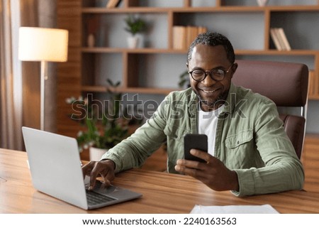 Glad busy handsome adult black businessman in glasses with computer typing on smartphone in home office interior. App for business, gadget for work, data analysis and customer support, covid-19 virus Royalty-Free Stock Photo #2240163563