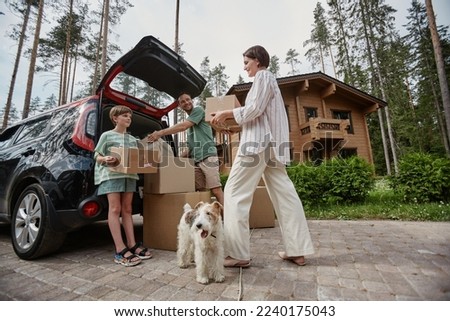 Full length portrait of happy family unloading boxes from car trunk while moving into new house with pet dog Royalty-Free Stock Photo #2240175043