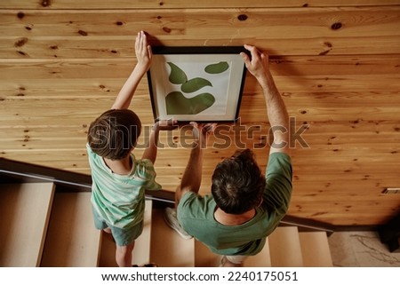 Top view of father and son hanging picture together on wooden wall in new house