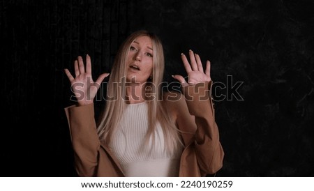 Portrait of a blonde woman in a business suit against a dark wall. The blonde is unhappy with the circumstances, makes a negative sign with her hands