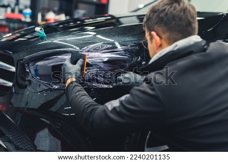Rear view of man squatting by black car to apply PPF paint protection film on headlight in a garage. Indoor horizontal shot. High quality photo