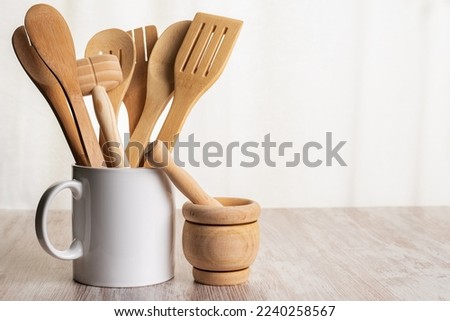 Wooden kitchen utensils such as spoons, forks and tongs inside a jar and next to a kitchen mortar on a wooden counter. Royalty-Free Stock Photo #2240258567