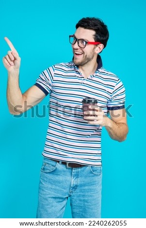 Caucasian Handsome Brunet Man With Brown Paper Cup While Posing in Glasses Gesturing With Point Finger Against Blue Background. Vertical Orientation