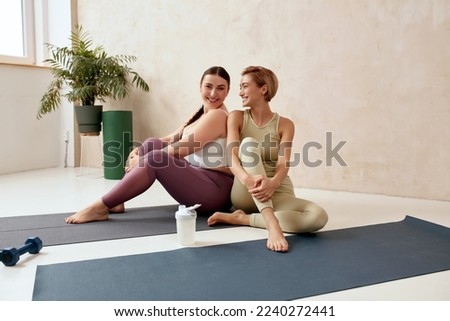 Smiling Woman Embracing After Yoga. Female Friends Posing After Training Together At Home. Attractive Trainer And Her Client In Sportswear Sitting On Mat Royalty-Free Stock Photo #2240272441