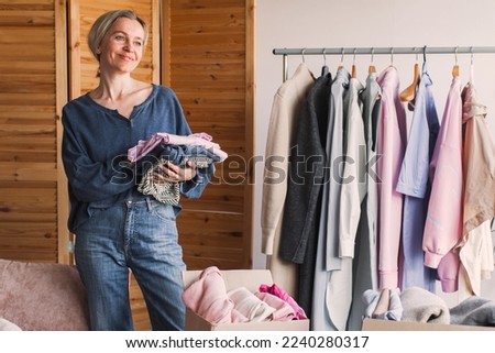 Happy middle aged woman holding cardboard donation box with clothes. Smiling compassionate female preparing staff for charity, reusage recycling concept.