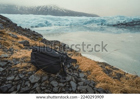 Close up black bag on pebble near lake concept photo. Front view photography with mountains and glacier on background. High quality picture for wallpaper, travel blog, magazine, article